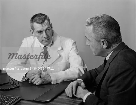 1950s DOCTOR SITTING AT DESK TALKING TO MALE PATIENT