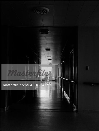 1960s - 1970s NURSE PUSHING ELDERLY FEMALE PATIENT DOWN DARK HOSPITAL HALLWAY IN WHEELCHAIR