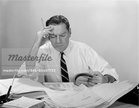 1960s BUSINESSMAN DESK FULL PAPERS HAND TO FOREHEAD SERIOUS EXPRESSION