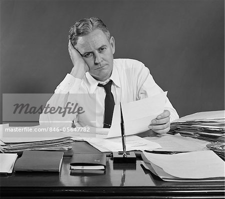 https://image1.masterfile.com/getImage/846-05647782em-1950s-unhappy-overworked-businessman-sitting-at-desk-holding-papers.jpg