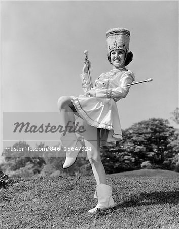 1960s SMILING MAJORETTE IN UNIFORM WITH SHORT SKIRT BOOTS TALL HAT AND TWO BATONS POSING OUTDOORS