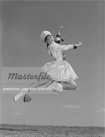 1960s WOMAN MAJORETTE WEARING BAND UNIFORM HOLDING BATON JUMPING INTO THE AIR