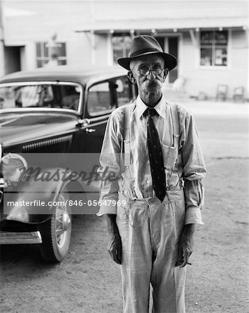1930s PORTRAIT OF OLD MAN WEARING HAT GLASSES TIE & SUSPENDERS CAR IN BACKGROUND