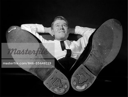 1960s BUSINESSMAN IN SHIRTSLEEVES HANDS BEHIND HEAD SITTING WITH SHOES ON DESK DAYDREAMING