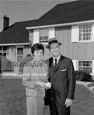 1960s COUPLE SMILING MAN AND WOMAN STANDING BEFORE SUBURBAN HOUSE