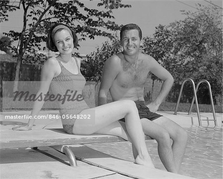 1960s MAN WOMAN COUPLE SITTING ON DIVING BOARD ON SIDE OF SWIMMING POOL