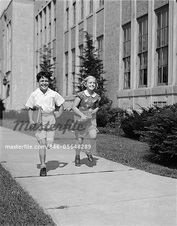 1940s BOY GIRL RUNNING DOWN SIDEWALK CARRYING SCHOOL BOOKS SMILING