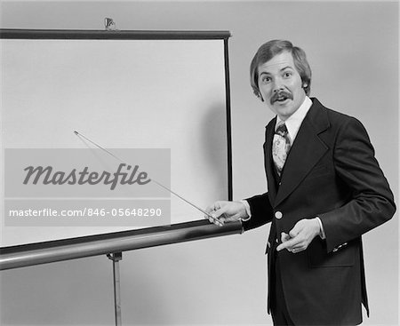 1970s YOUNG MAN SPEAKING POINTING TO BLANK SLIDE SCREEN FOR PRESENTATION