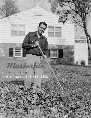 1950s - 1960s SMILING MAN RAKING AUTUMN LEAVES IN FRONT YARD OF HOUSE