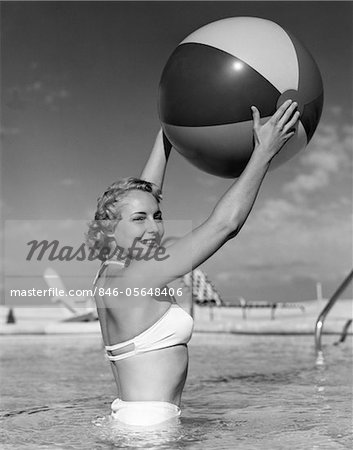 1950s SIDE VIEW OF BLONDE IN WHITE BIKINI STANDING IN POOL IN WAIST-HIGH WATER HOLDING BEACH BALL IN AIR