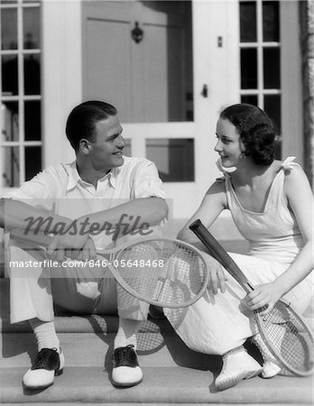 1930s COUPLE MAN WOMAN IN TENNIS WHITES HOLDING RACQUETS SITTING TALKING ON STEPS IN FRONT OF HOUSE OUTDOOR