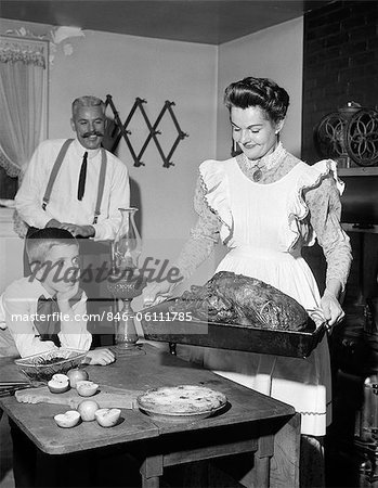 1950s MOCK TURN OF THE 20TH CENTURY THANKSGIVING DINNER SMILING MOTHER SHOWING ROAST TURKEY TO PLEASED HUSBAND AND SURPRISED SON