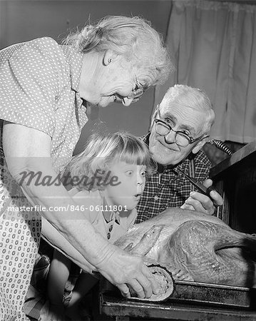 1950s 1960s EXCITED LITTLE GIRL GRANDDAUGHTER WITH GRANDPARENTS WATCHING ROAST TURKEY COMING OUT OF OVEN