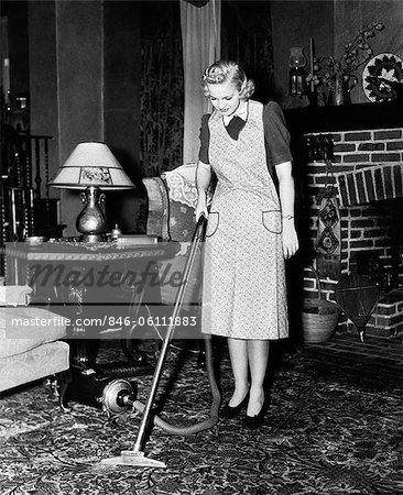 1930s 1940s WOMAN HOUSEWIFE WEARING APRON PUSHING ELECTRIC VACUUM CLEANER IN ORNATE LIVING ROOM HOUSE CHORE