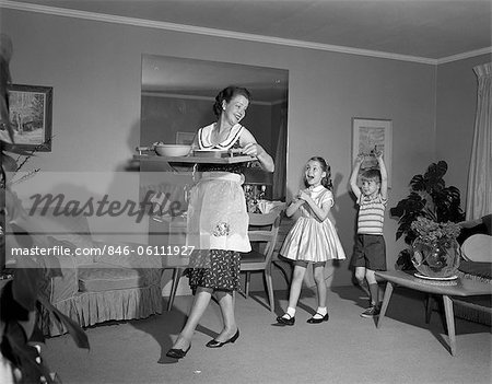 1960s MOTHER WALKING WITH TRAY OF SNACK FOOD HAPPY SON AND DAUGHTER FOLLOWING HER INDOOR