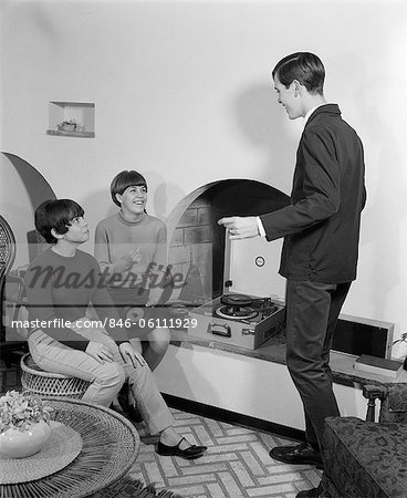 1960s 2 TEEN GIRLS SEATED ON WICKER STOOL AT FIREPLACE CHANGING 45 RPM RECORDS WITH TEEN BOY DANCING