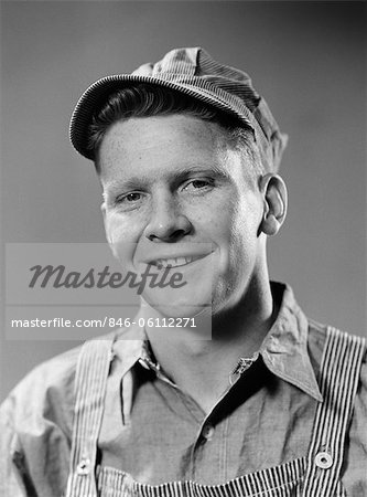 1940s PORTRAIT OF SMILING BLUE COLLAR WORKING MAN WEARING OVERALLS AND ENGINEER HAT LOOKING AT CAMERA
