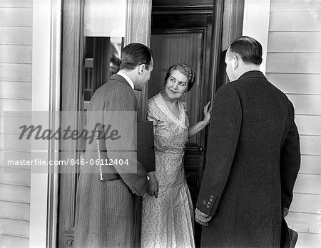 1930s 1940s TWO DOOR-TO-DOOR SALES SALESMEN TALKING TO HOUSEWIFE AT FRONT DOOR MAKING SALES PRESENTATION