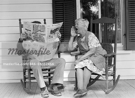 1970s ELDERLY COUPLE IN ROCKING CHAIRS ON PORCH MAN READING NEWSPAPER WHILE WIFE HOLLERS AT HIM