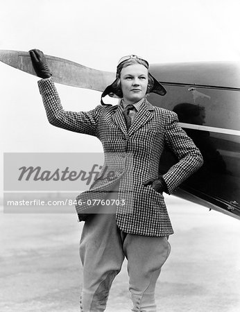 1930s WOMAN AVIATOR PILOT STANDING NEXT TO AIRPLANE ONE HAND ON PROPELLER FLIGHT GOGGLES CAP TWEED SUIT FASHION