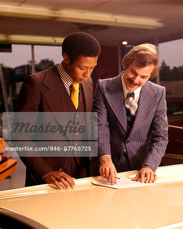 1970s TWO MEN CUSTOMER AND SALESMAN READING SALES CONTRACT ON HOOD OF NEW CAR IN DEALERSHIP SALES SHOWROOM