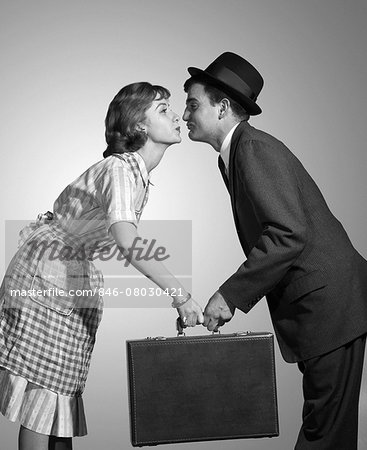 1950s 1960s HOMEMAKER WIFE IN CHECKED APRON KISSING BUSINESSMAN HUSBAND IN SUIT HAT AND TIE AS SHE HANDS HIM A BRIEFCASE