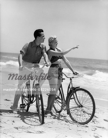 1930s COUPLE MAN WOMAN BIKING ON BEACH WOMAN POINTING TO HORIZON