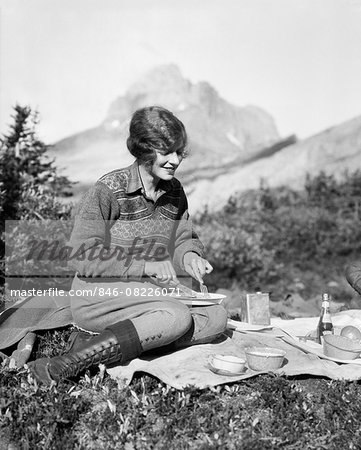 1920s WOMAN CAMPING EATING BREAKFAST OUTDOORS SITTING ON GRASS MOUNTAINS IN BACKGROUND BAKER LAKE ALBERTA CANADA