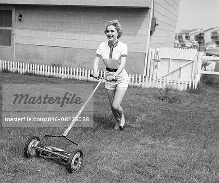 1950s SMILING BLOND WOMAN IN SHORTS MOWING LAWN WITH PUSH MOWER