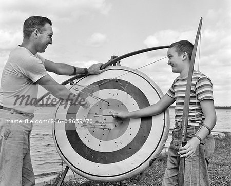 1950s SMILING FATHER AND SON BY LAKE PULLING ARROWS OUT OF ARCHERY TARGET BULL'S EYE