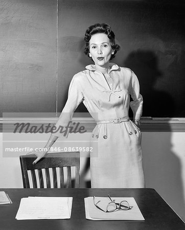 1950s WOMAN SCHOOL TEACHER LOOKING AT CAMERA STANDING IN FRONT OF CHALKBOARD IN CLASSROOM LEANING ON CHAIR BY DESK