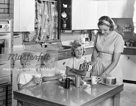 1950s MOTHER THREE DAUGHTERS STANDING AROUND KITCHEN TABLE MAKING LUNCH PEANUT BUTTER JELLY SANDWICHES MOTHER POURING GLASS MILK