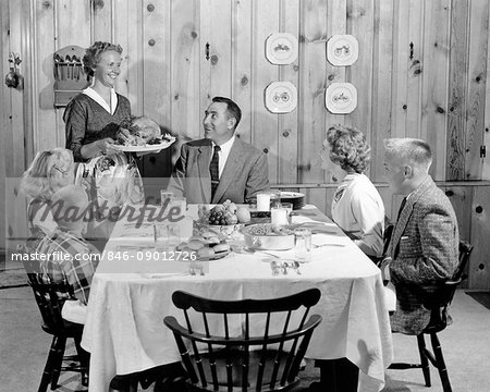 1950s MOTHER SERVING TURKEY ON PLATTER TO SMILING FAMILY AT TABLE IN KNOTTY PINE PANELED DINING ROOM