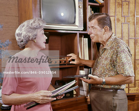 1960s SENIOR COUPLE LISTENING TO RECORDED MUSIC MAN TURNING STEREO PHONOGRAPH DIAL WOMAN HOLDING LP RECORD ALBUM COVERS 1960s SENIOR COUPLE LISTENING TO RECORDED MUSIC MAN TURNING STEREO PHONOGRAPH DIAL WOMAN HOLDING LP RECORD ALBUM COVERS