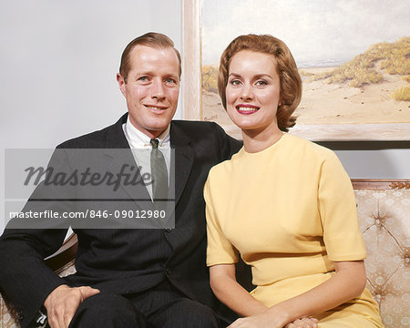 1960s HAPPY COUPLE SITTING ON SOFA MAN WEARING BUSINESS SUIT TIE WOMAN IN A YELLOW DRESS LOOKING AT CAMERA SMILING