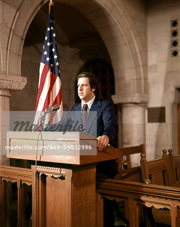 1970s MAN STANDING SPEAKING AT PODIUM BESIDE AN AMERICAN FLAG