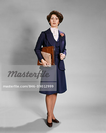 1970s BUSINESSWOMAN STANDING LOOKING AT CAMERA WITH SERIOUS FACIAL EXPRESSION HOLDING NEWSPAPER AND BRIEFCASE WEARING BLUE SUIT