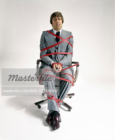 1970s BUSINESS MAN TIED UP IN RED TAPE SITTING IN OFFICE SWIVEL CHAIR