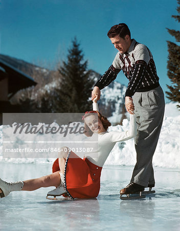 1940s 1950s COUPLE ICE SKATING SMILING WOMAN LOOKING AT CAMERA HAS SLIPPED AND FALLEN MAN HOLDING HER HANDS IS LIFTING HER UP