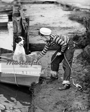 1940s TEENAGE BOY WEARING SAILOR HAT STRIPED SHIRT BLUE JEANS LANDING ROW BOAT CARRYING PET DOG