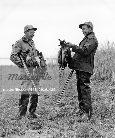 1950s TWO MEN DUCK HUNTERS WITH SHOTGUNS ADMIRING A BRACE OF DUCKS