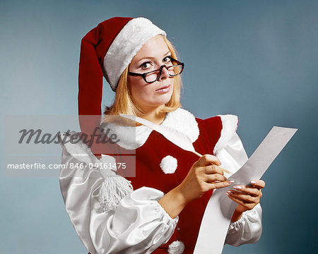 1960s WOMAN IN CHRISTMAS SANTA'S HELPER COSTUME AND CAP WEARING GLASSES LOOKING AT CAMERA CHECKING NAUGHTY OR NICE LIST