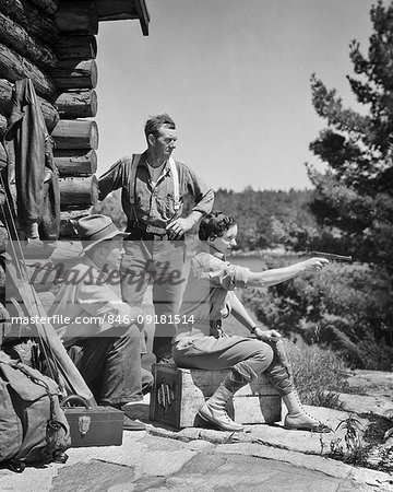1930s TWO MEN CAMP GUIDES WATCHING INSTRUCTING YOUNG WOMAN SHOOTING 22 CALIBER PISTOL OUTSIDE RUSTIC MOUNTAIN LOG CABIN
