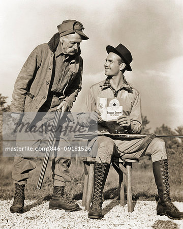 1930s 1940s OLD HUNTER HOLDING SHOTGUN SPEAKING TO SMILING YOUNGER MAN READING STRAIGHT SHOOTING INSTRUCTIONS HOLDING 22 RIFLE