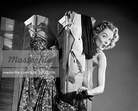 1950s FLIRTING WOMAN LOOKING AT CAMERA PEEKING AROUND FROM BEHIND DRESSING SCREEN HUNG WITH DRESS BRA AND SLIP