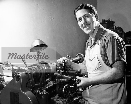 1950s PORTRAIT SMILING MAN FACTORY WORKER IN APRON WORKING WITH METAL ON LATHE LOOKING AT CAMERA