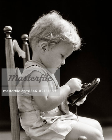 1930s TWO YEAR OLD BLOND BOY SITTING IN CHAIR CONCENTRATING ON HOW TO UNTIE A SHOE LACE