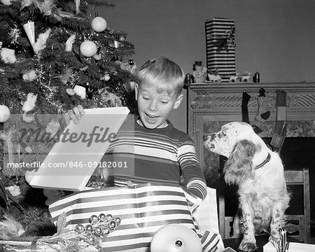 1950s SURPRISED BOY OPENING CHRISTMAS PRESENT WITH DOG WATCHING BY TREE