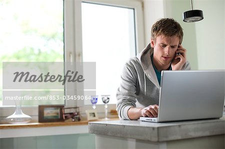 Young man standing at a counter and using mobile phone and laptop, low angle view