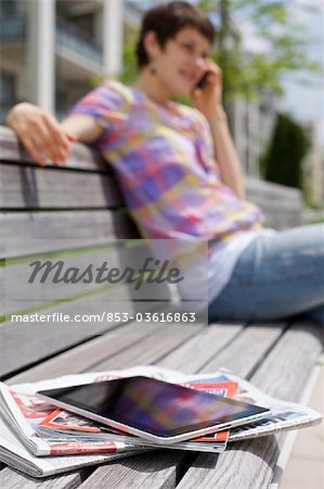 Young woman with cell phone, iPad, newspapers and magazine on a bench